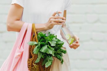 Hands holding bags, one which contains a green leafed vegetable, whilst also holding a water bottle in the other hand